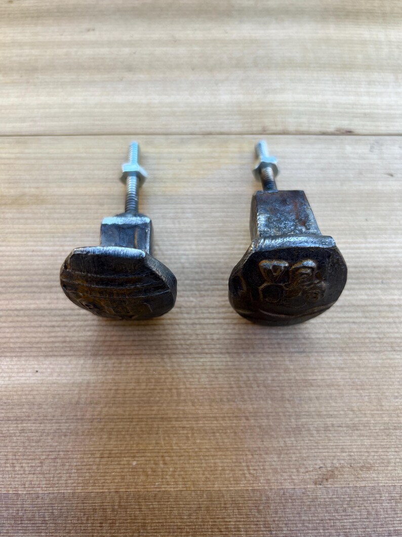 May include: Two weathered, dark metal cabinet knobs with an oval design. Each knob has a threaded bolt and nut for attachment. The knobs are set against a light brown wooden surface.