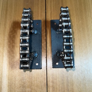 May include: Two industrial-style door handles made from metal chain links and mounted on black rectangular plates. The handles are a dark metallic color, and the plates have screw holes for mounting. The background is a light brown wooden surface.