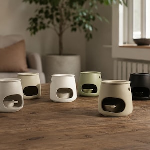 May include: A collection of six ceramic wax warmers in various matte colors including white, cream, sage green, and black. Each warmer features a rounded, tapered body with a wide front opening and a small circular vent hole on the side.