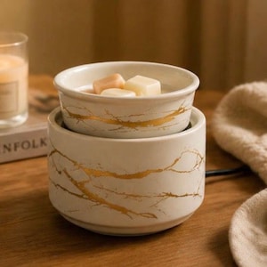 May include: A white ceramic wax warmer featuring a gold marble-like vein pattern. The cylindrical base supports a removable bowl containing three solid wax melts. The design is consistent across both the base and the bowl, creating a cohesive look.