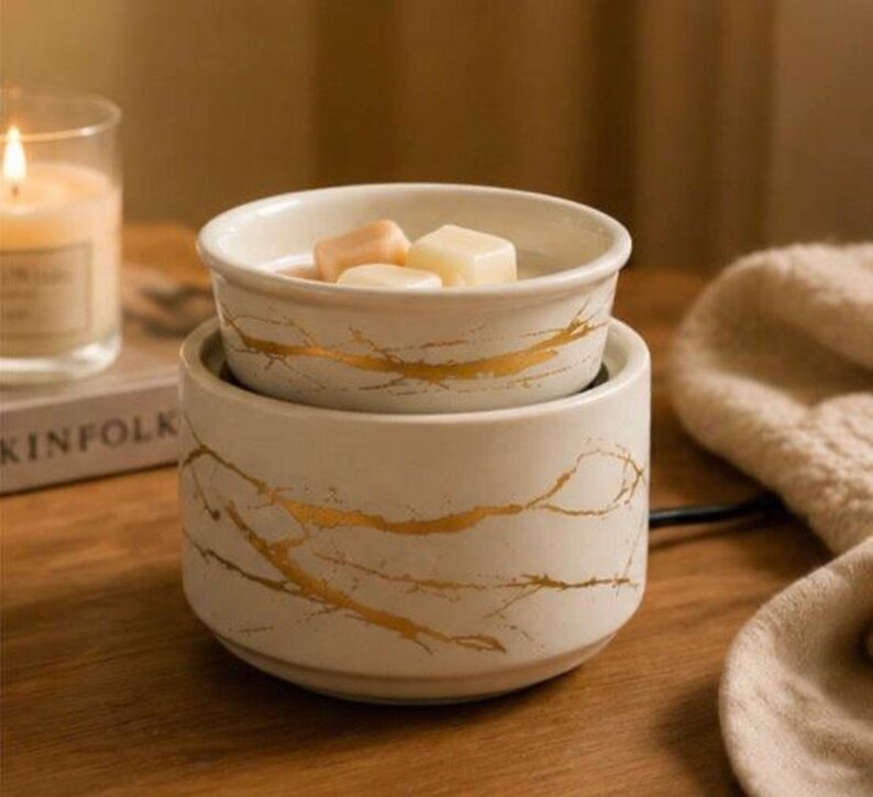 May include: A white ceramic wax warmer featuring a gold marble-like vein pattern. The cylindrical base supports a removable bowl containing three solid wax melts. The design is consistent across both the base and the bowl, creating a cohesive look.