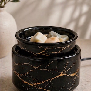 May include: A black ceramic wax warmer with a gold-colored marble-like vein pattern. The cylindrical base supports a removable bowl containing several solid, multi-colored wax melts in shades of cream, light gray, and soft peach.
