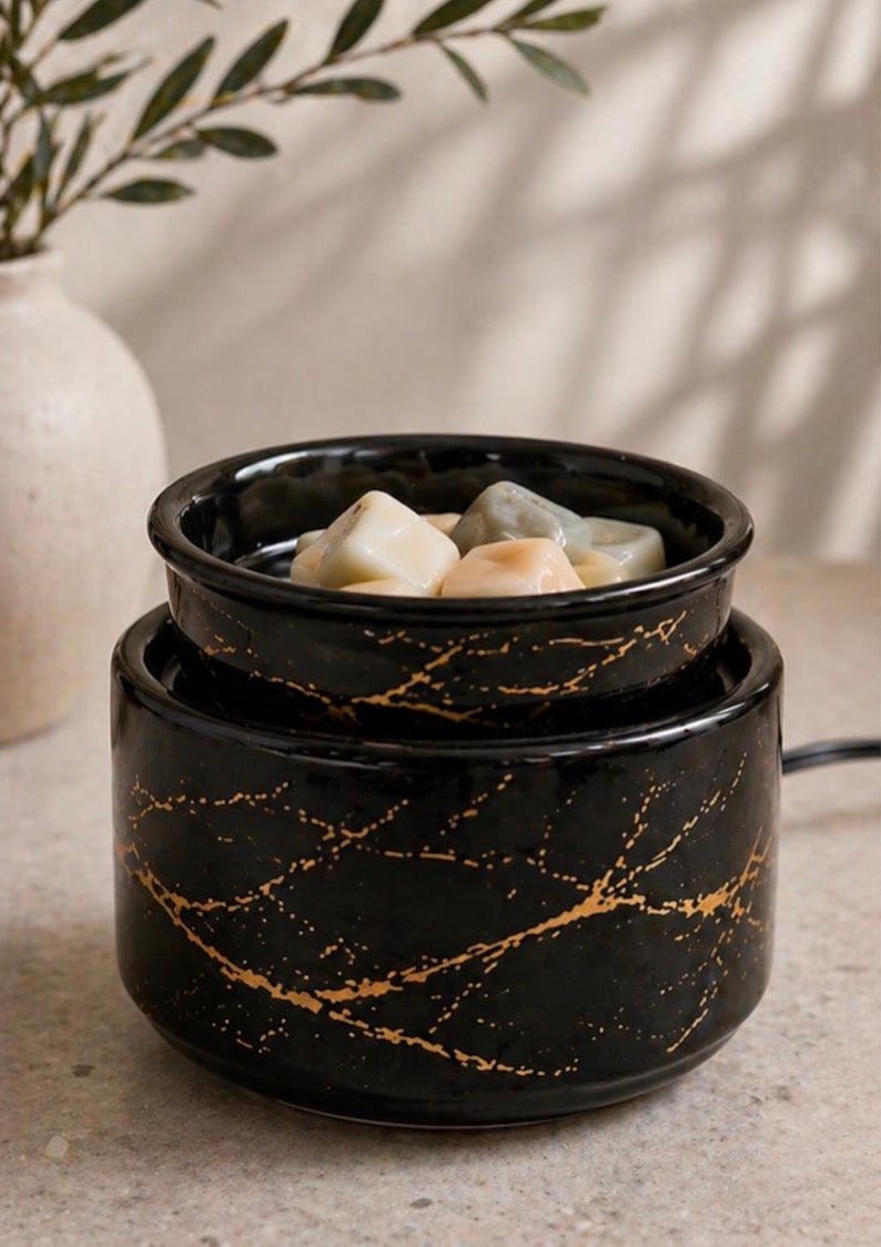 May include: A black ceramic wax warmer with a gold-colored marble-like vein pattern. The cylindrical base supports a removable bowl containing several solid, multi-colored wax melts in shades of cream, light gray, and soft peach.