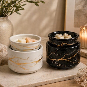 May include: Two ceramic wax warmers, one white and one black, both featuring a gold marbled vein pattern. Each warmer consists of a cylindrical base and a removable top dish containing small, light-colored wax melts for fragrance diffusion.