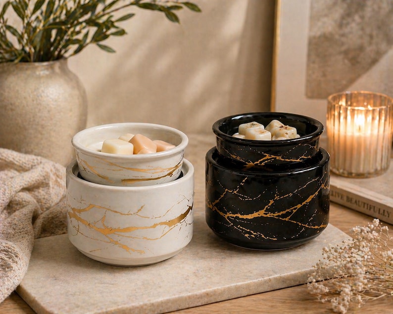 May include: Two ceramic wax warmers, one white and one black, both featuring a gold marbled vein pattern. Each warmer consists of a cylindrical base and a removable top dish containing small, light-colored wax melts for fragrance diffusion.