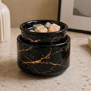 May include: A black ceramic wax warmer with a gold marble-like vein pattern. The cylindrical base supports a removable top dish containing several small, solid, multi-colored wax cubes in shades of light gray, beige, and soft peach.