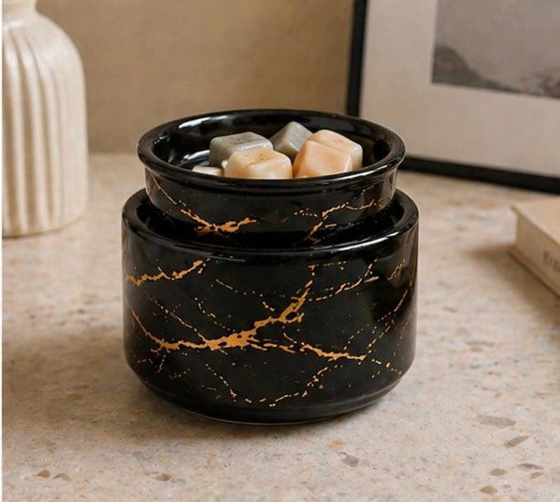 May include: A black ceramic wax warmer with a gold marble-like vein pattern. The cylindrical base supports a removable top dish containing several small, solid, multi-colored wax cubes in shades of light gray, beige, and soft peach.