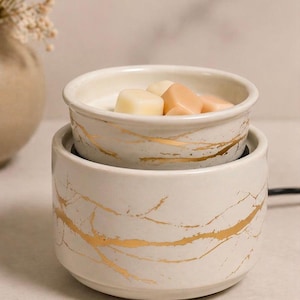 May include: A cylindrical ceramic wax warmer featuring a cream-colored base with a gold-toned marbled pattern. The top section is a removable dish containing several small, solid, light-colored wax cubes intended for melting.