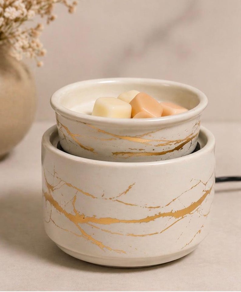May include: A cylindrical ceramic wax warmer featuring a cream-colored base with a gold-toned marbled pattern. The top section is a removable dish containing several small, solid, light-colored wax cubes intended for melting.
