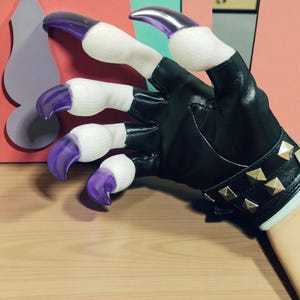 Op de afbeelding: Een zwarte imitatielederen handschoen met zilveren piramidevormige studs op de pols. De handschoen heeft witte vingerbedekkingen en paarse klauw-vormige vingernagels. De handschoen ligt op een houten oppervlak.