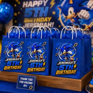 May include: Blue gift bags with white handles feature Sonic the Hedgehog graphics and the text "Jeremiah's 5th Birthday." The bags are arranged on a wooden display with a sign that reads "Thank you for racing by!"