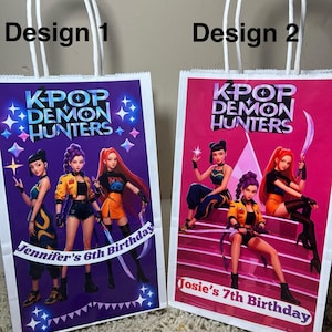May include: Two white paper gift bags with white handles. Each bag features a colorful graphic of anime-style characters with the text "K-POP DEMON HUNTERS." One bag says "Jennifer's 6th Birthday," the other says "Josie's 7th Birthday."
