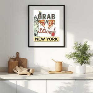 Könnte beinhalten: Ein gerahmter Druck mit einem Retro-Design, der den Text "GRAB A SLICE NEW YORK" zeigt. Das Kunstwerk enthält Illustrationen von Pizzastücken, der Freiheitsstatue und einem Getränk. Der Rahmen ist schwarz.
