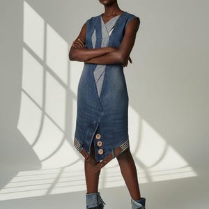 Könnte beinhalten: Ein ärmelloses Jeanskleid mit V-Ausschnitt und Wickeldesign. Das Kleid hat Holzknöpfe, braune Lederakzente und wird mit passenden Jeans-High-Heel-Stiefeln kombiniert. Das Model steht vor einem weißen Hintergrund.