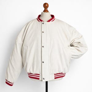 Könnte beinhalten: Cremefarbene Cordjacke mit rot-weiß gestreiftem Kragen, Bündchen und Saum. Die Jacke hat eine Knopfleiste vorne und zwei Seitentaschen. Die Jacke wird auf einer Schaufensterpuppe präsentiert.