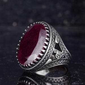 May include: A silver ring featuring a large, oval-cut, deep red gemstone. The ring has a detailed, braided pattern on the band and a textured setting. The gemstone is faceted, catching the light.