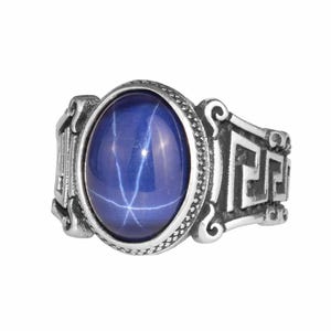 May include: A silver ring featuring a large, oval, deep blue gemstone with a star-like pattern. The ring has a decorative band with a Greek key design and scrollwork on the sides. The gemstone is set in a textured bezel.