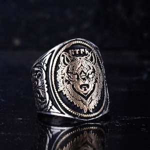 以下が含まれることがあります： 金と黒の狼の頭のデザインが施されたシルバーリング。リングには複雑な模様と上部にテキストが施されています。狼は楕円形の中央に配置され、背景は暗いです。
