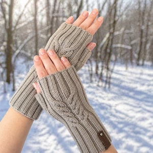 May include: A pair of light brown fingerless gloves made of knitted material. The gloves have a cable knit design on the back of the hand and a small brown leather tag. The background is a snowy winter scene.