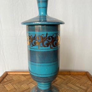 Könnte beinhalten: Ein türkisfarbenes und dunkelblaues Keramikgefäß mit Deckel und Sockel. Das Gefäß hat goldene Schnörkel und horizontale Streifen. Der Deckel hat einen Griff. Das Gefäß steht auf einer gewebten Oberfläche.