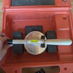 May include: A precision angle level tool with a circular dial and a horizontal spirit level vial, featuring a beige handle and black end cap. The tool is housed in a bright orange plastic carrying case with dark grey foam inserts for protection.