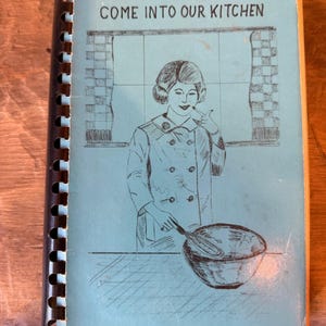 May include: A vintage cookbook with a light blue cover and spiral binding. The cover features the text "COME INTO OUR KITCHEN" above an illustration of a woman in a chef's coat holding a whisk over a bowl.