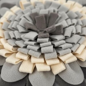 May include: Close-up of a multi-layered, flower-shaped pet bed in shades of gray, cream, and dark gray. The petals are made of soft, folded fabric, creating a textured, plush appearance. The design is centered, with a gradient effect from the center to the outer edges.