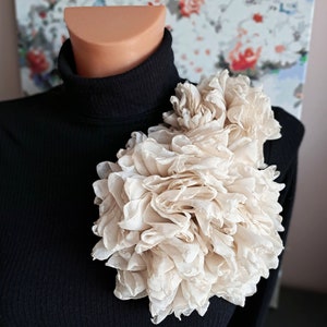Puede incluir: Un jersey de cuello alto negro adornado con una gran flor de tela en capas de color crema. La flor es el punto focal, con su delicada textura arrugada. El fondo presenta una colorida pintura abstracta.