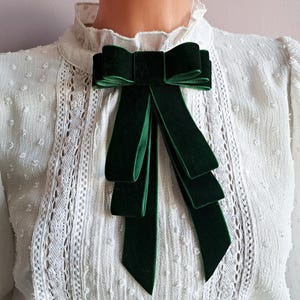 May include: A dark green velvet bow tie with long, cascading ribbons. The bow is centered on a white, textured blouse with a high, ruffled collar and lace detailing. The bow tie adds a touch of elegance.