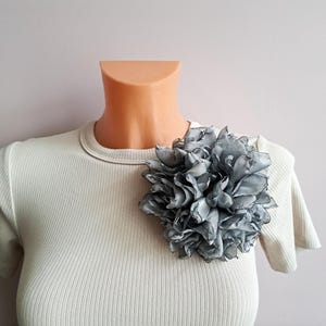 Puede incluir: Un broche de flor de tela gris prendido a una camiseta crema acanalada. La flor es grande y tiene múltiples capas de pétalos. La camiseta tiene mangas cortas y cuello redondo. El broche está sujeto a la camiseta en un maniquí.