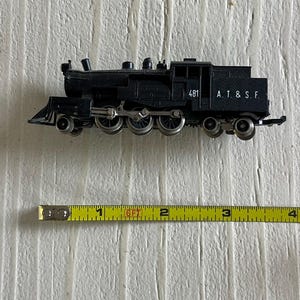 May include: A black model train with the text "481 A.T. & S.F." is displayed next to a yellow measuring tape. The train has multiple wheels and detailed features. The measuring tape shows a length of approximately 4 inches.