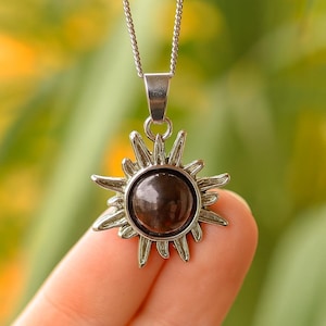 Pode incluir: Um colar com pingente prateado com um design de sol. O pingente tem uma pedra circular marrom escura no centro. O sol tem raios pontiagudos ao redor da pedra. A corrente do colar também é prateada.