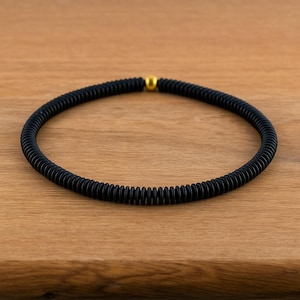 May include: A black, circular necklace with a textured, ribbed design. The necklace features a small, gold-colored bead at the center. The jewelry is displayed on a light brown wooden surface.