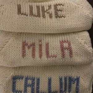 May include: Three cream-colored, knitted sweaters stacked on top of each other. Each sweater has a different name embroidered across the front in a contrasting color: "LUKE" in brown, "MILA" in pink, and "CALLUM" in blue. The sweaters have a crew neck design.