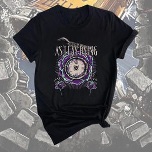 Puede incluir: Camiseta negra con el texto "AS I LAY DYING" sobre un diseño floral morado y blanco con una esfera de reloj. Un pájaro negro está posado sobre el texto. Dos rosas moradas están debajo.