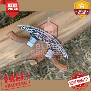 Puede incluir: Un hacha artesanal con una hoja curva con un diseño en blanco y negro. El hacha tiene un mango de madera y el texto "BLADE BRIGADE" es visible. El hacha se muestra sobre una superficie de madera.