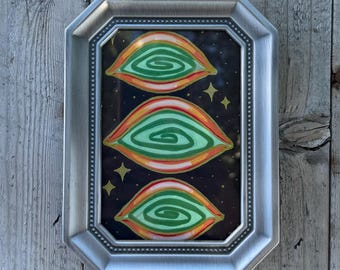 May include: A framed artwork featuring three stylized eye shapes in green, orange, and red, set against a black background with gold star accents. The artwork is encased in a silver-colored, octagonal frame.