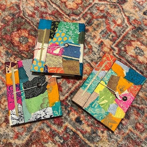 May include: Three handmade journals with colorful patchwork covers. Each journal features a unique combination of patterned fabrics in shades of blue, green, orange, and pink. The journals are bound with string and button closures.