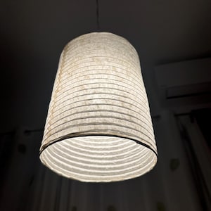May include: A hanging pendant light with a cylindrical lampshade. The lampshade is made of a textured, off-white material with horizontal lines. The light is suspended from a thin wire and illuminates the room below.