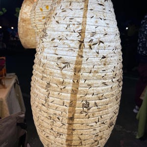 Puede incluir: Un grupo de faroles de papel colgantes con un brillo cálido y acogedor. Los faroles tienen forma cilíndrica, con una superficie texturizada de color blanquecino incrustada con pequeños detalles oscuros en forma de hoja. Una franja vertical marrón recorre el centro de cada farol.