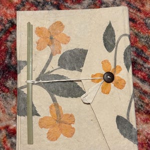 Op de afbeelding: Een handgemaakt notitieboek met een bloemenontwerp. De kaft is lichtbeige met geperste oranje bloemen en groene bladeren. Een dunne groene stok en een touwsluiting bevinden zich aan de linkerkant. Een donkerbruine knoop bevindt zich aan de rechterkant.