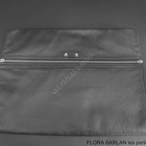 Peut inclure: Pochette en cuir noir avec une fermeture éclair argentée et une petite boucle sur le côté. La pochette est de forme rectangulaire et présente une surface texturée. Le texte "LES PERLES D'ANTAN" est visible.