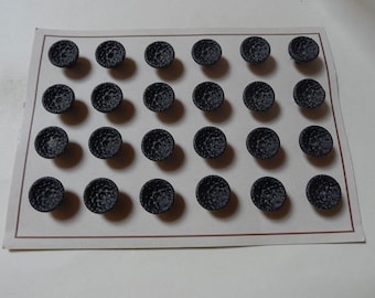 May include: Twenty-four dark grey buttons with a textured, dimpled surface are arranged on a rectangular card. The buttons are evenly spaced and appear to be made of a solid material. The card has a thin, brown border.