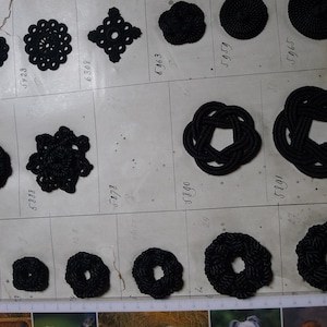 Puede incluir: Una colección de varios adornos decorativos negros, que incluyen rosetas, nudos y diseños circulares. Los adornos están dispuestos en una cuadrícula, con medidas en centímetros en la parte inferior. Probablemente utilizados para manualidades o moda.