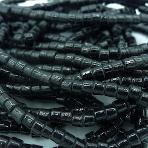 May include: Close-up of a pile of black, square-shaped beads. The beads are strung together, creating a textured and shiny surface. The beads are likely used for jewelry making or other crafts.