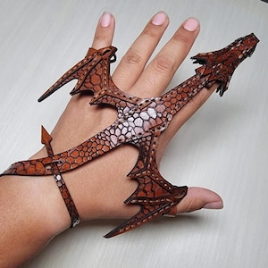 May include: A brown leather dragon claw hand accessory. The dragon claw is made of brown leather with a scale pattern and is designed to be worn on the fingers and wrist. The dragon claw has a detailed design with sharp edges and a textured surface.