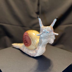 May include: A ceramic snail figurine with a cream-colored body and a shell that is yellow and red. The snail has two antennae with black tips and is posed on a dark surface.