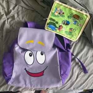 Puede incluir: Una mochila morada con un diseño de cara sonriente y un mapa a juego. La mochila tiene una solapa con detalles amarillos y dos bolsillos laterales. El mapa presenta personajes de dibujos animados y un paisaje colorido.