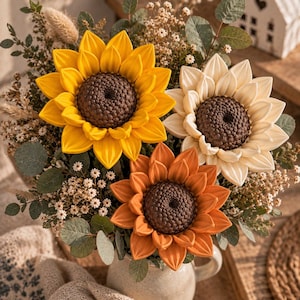 May include: A bouquet of three large artificial sunflowers in yellow, cream, and burnt orange, each featuring a dark brown textured center. The flowers are arranged with dried greenery, small white filler flowers, and eucalyptus leaves in a ceramic pitcher.