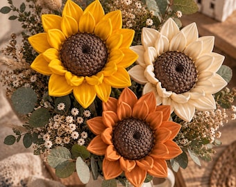 May include: A bouquet of three large artificial sunflowers in yellow, cream, and burnt orange, each featuring a dark brown textured center. The flowers are arranged with dried greenery, small white filler flowers, and eucalyptus leaves in a ceramic pitcher.